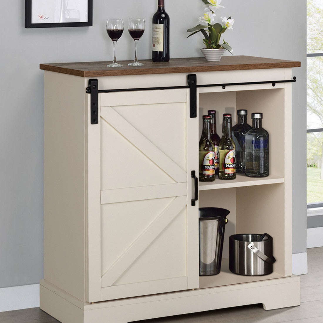 A beige farmhouse sideboard with a sliding barn door, featuring open shelving, and arranged with bottles and a ice bucket on the lower shelf.