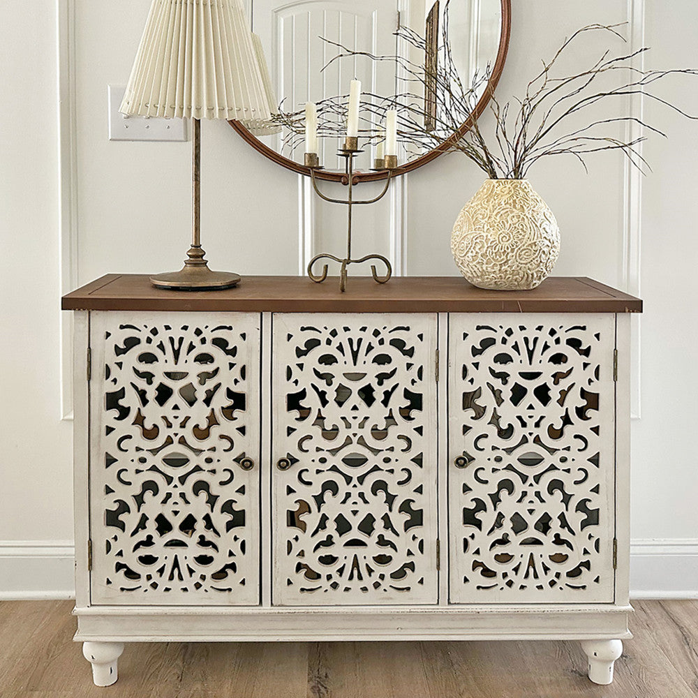 A vintage white storage cabinet with intricate carved designs on the doors, featuring a wooden top. The cabinet is placed in a bright room and is used to store items, providing a practical storage solution in a stylish manner.