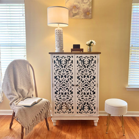 A tall accent storage cabinet with a decorative carved design in a room setting, featuring a lamp and decorative items on its top.